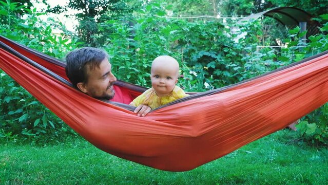 Portrait of curious cute little toddler child daughter with her father resting in hammock looking at camera. Family at backyard garden in summer sunny day.