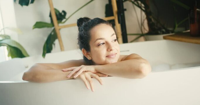 Young woman rests in bathtub putting arms on bath edge