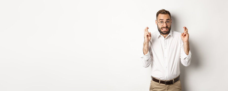 Worried man making a wish, cross fingers and hope for relish, standing over white background