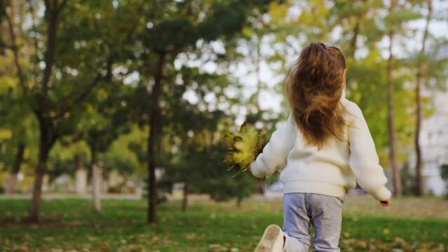 Happy girl runs in autumn park with bouquet of yellow leaves in her hand. Child plays, runs cheerfully on warm autumn day. Concept of healthy childhood and happy family. Autumn walk, daughter's smile