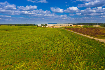 Równina pokryta łąkami i polami uprawnymi przecinanymi przez drogę. W oddali widać plac budowy, konstrukcje dużych hal. Zdjęcie z drona.