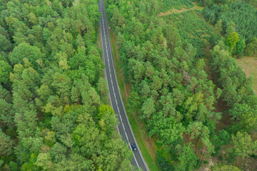 Asfaltowa droga w lesie. Widoczne są białe linie na jezdni. Pobocza porasta zielona trawa, w głębi zielony las. Zdjęcie wykonane z użyciem drona.