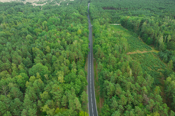 Asfaltowa droga w lesie. Widoczne są białe linie na jezdni. Pobocza porasta zielona trawa, w głębi zielony las. Zdjęcie wykonane z użyciem drona.