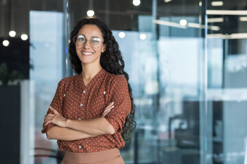Young beautiful and successful hispanic businesswoman smiling and looking at camera, female worker working inside office wearing glasses and curly hair, arms crossed portrait