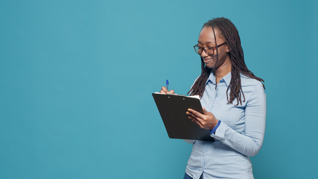 African american woman writing text on papers, using files on clipboard to take notes over blue background. Person putting information on documents and paperwork, feeling cheerful.