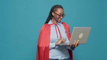 Woman dressed as superhero using laptop in studio, wearing red cape hero costume and browsing internet. Looking at computer with online network, having cloak and cartoon comic look. © DC Studio