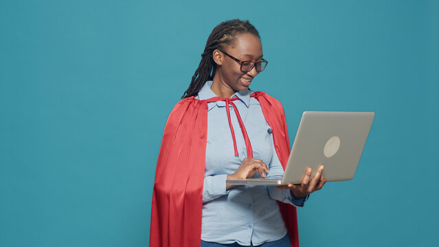 Woman dressed as superhero using laptop in studio, wearing red cape hero costume and browsing internet. Looking at computer with online network, having cloak and cartoon comic look.
