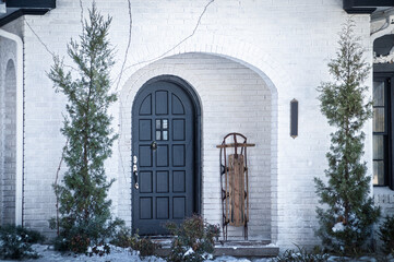 Snow Day - Porch of white painted brick house with arched front door and sled propped up nearby -...