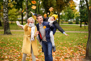 Happy mom, dad and son having fun together in the autumn park, throwing golden leaves and laughing.