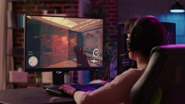 Over shoulder view of gamer girl using pc gaming setup relaxing playing multiplayer online action video game talking using headset. Woman streaming first person shooter while explaining gameplay.