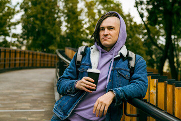 Young handsome man with dreadlocks in casual clothes drinking coffee from paper cup standing on footbridge in city park.