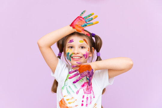 A beautiful little girl stained in multicolored paints on a pink isolated background has fun smiling.