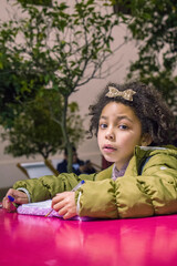 curly latin little girl wearing a winter jacket is looking at camera while writing on her personal notebook at the table outdoors. copy space, vertical.