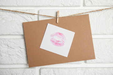 Envelope and card with lip print hanging on twine near white brick wall. Love letter