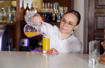 Young waitress in glasses standing at bar, pouring juice into glass from jug in restaurant.