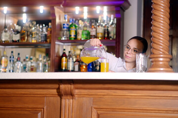 Young waitress in glasses standing at bar, pouring juice into glass from jug in restaurant.