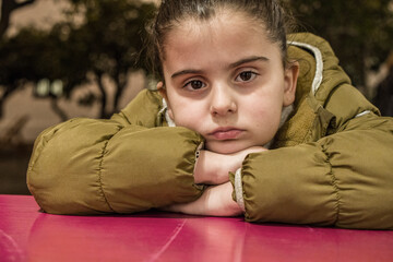 Unhappy little girl portrait with boring expression leaning on table outdoors wearing a winter jacket.