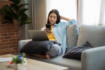 Fototapeta premium Young asian woman smiling and using smartphone on couch at home.