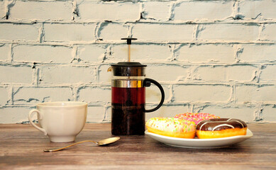 A plate with three donuts in multi-colored glaze, a teapot and a cup of hot tea on a wooden table.