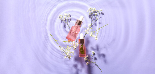 Bottles of cosmetic serum and flowers in water on lilac background, top view