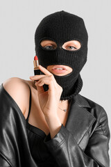 Young woman in balaclava holding red lipstick on light background