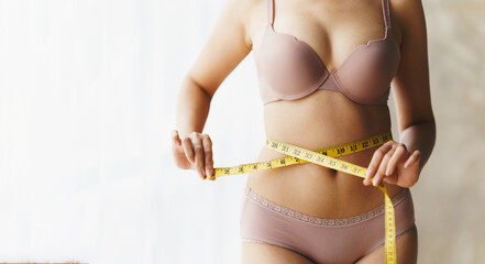 A woman wearing a bra is using a waist tape to measure her waist size.