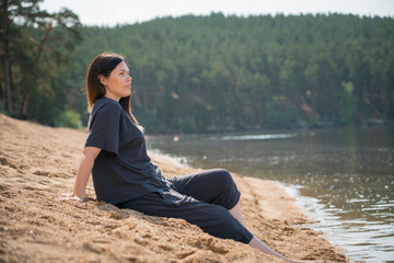 Naklejka na ściany i meble A young pregnant woman sits on the beach near the water and admires the view. Unity with nature, alone with yourself, grounding, harmony, stress relief, relaxation, mental healing. High quality photo