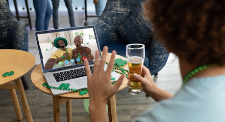 Mixed race man holding beer at bar making st patrick's day video call waving to couple on laptop - Powered by Adobe