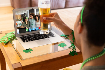 Mixed race woman holding beer having st patrick's day video call with friends on laptop at home