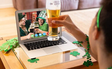 Mixed race woman holding beer having st patrick's day video call with friends on laptop at home
