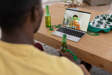 African american man holding beer making st patrick's day video call to friend on laptop at home