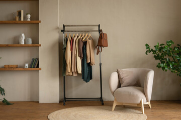 Rail rack with a capsule wardrobe in the living room against a beige wall. Trendy outfits in autumn colors: burgundy, beige, khaki, green, brown.