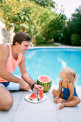 Dad cuts watermelon into slices in front of a little girl sitting by the pool. High quality photo