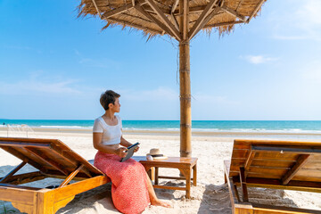 Modern Asian businesswoman working on digital tablet with internet at tropical beach in summer...