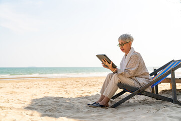 Modern Asian senior businesswoman working on digital tablet with internet at tropical beach in sunny day. Elderly retired woman enjoy outdoor lifestyle with using technology on summer holiday vacation