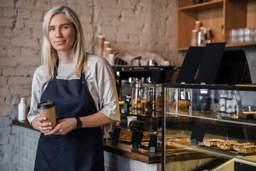 Shot of elderly female barista with coffee cup and apron in modern coffee shop.