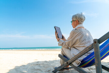 Modern Asian senior businesswoman working on digital tablet with internet at tropical beach in...