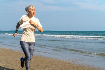 Happy Asian senior woman in sportswear jogging exercise on tropical beach in summer morning....