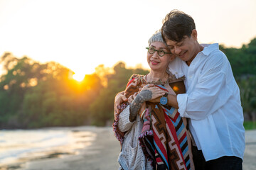 Happy Asian family senior couple enjoy outdoor lifestyle at the beach on summer holiday vacation. Senior man husband cover his wife with a blanket and hugging together on the beach at summer sunset
