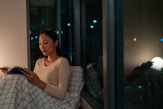  Young Asian woman sitting on the bed by the window in bedroom reading sad novel with crying. Attractive girl relax and enjoy indoors lifestyle leisure activity reading book before sleep at night.