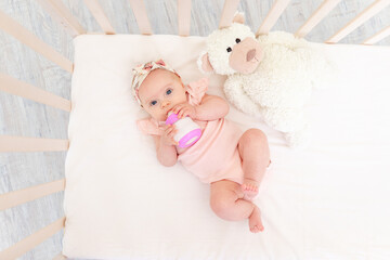 baby girl sucks a bottle of milk in the crib lying on her back with a toy in the nursery, baby food...