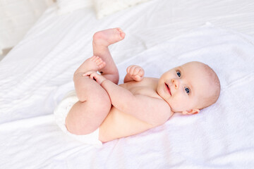 happy and laughing baby playing feet in diapers in a crib on a white cotton bed, hygiene and...