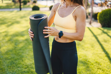 Woman's hands rolling yoga mat after training on a green meadow on a summer sunny day. Young active woman. Healthy lifestyle, sport. Fitness workout. Physical