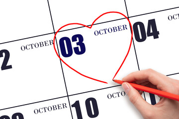 A woman's hand drawing a red heart shape on the calendar date of 3 October. Heart as a symbol of...