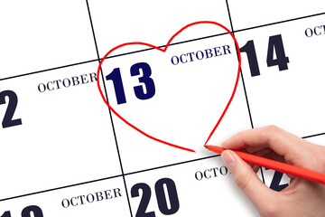 A woman's hand drawing a red heart shape on the calendar date of 13 October. Heart as a symbol of...