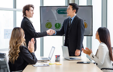Millennial Asian successful professional male businessmen in formal suit standing shaking hands together while unrecognizable female colleagues clapping hands applaud in company office meeting room