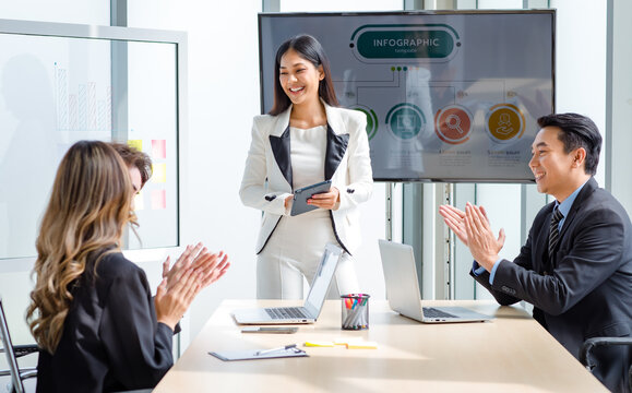 Millennial Asian successful professional businesswoman presenter speaker in white formal suit standing smiling holding tablet while male and female colleagues clapping hands applaud in meeting room