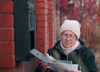 An elderly woman opens a mailbox on the fence and reads the correspondence she received. A moment in the life of a pensioner.