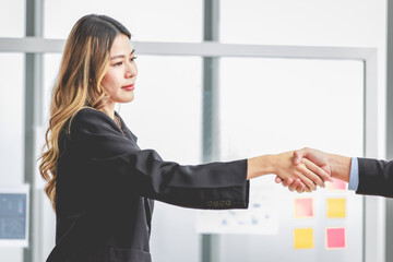 Millennial Asian successful professional female businesswoman in formal suit stand smiling shaking hands greeting with unrecognizable male businessman after agreement achievement deal done in office