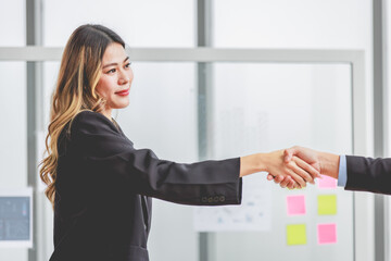 Millennial Asian successful professional female businesswoman in formal suit stand smiling shaking hands greeting with unrecognizable male businessman after agreement achievement deal done in office